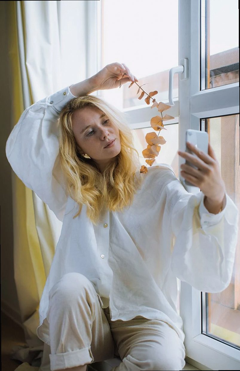 Blonde woman sitting by window, taking a selfie with a smartphone, enjoying a calm moment indoors.
